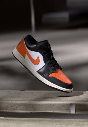 Zapatilla Nike negra, blanca y naranja flotando sobre un banco de hormigón con un fondo oscuro desenfocado.