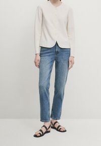 Cream button-up cardigan with a rounded hem, paired with blue straight-leg jeans and black strappy sandals. Simple design and textured fabric.