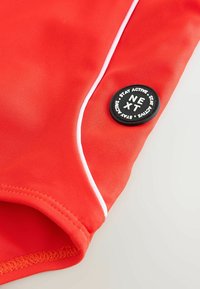 Rote sportliche Badehorts aus elastischem Material, mit weißen Paspeln und einem runden schwarzen Patch mit weißem Text "NEXT" und "STAY ACTIVE".