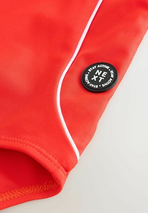 Červené sportovní plavecké šortky vyrobené z elastického materiálu, s bílými pruhy a kulatou černou nášivkou s bílým textem ‘NEXT’ a ‘STAY ACTIVE’.