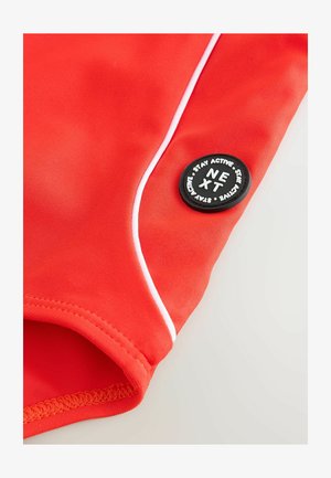 Shorts de bain de sport rouges en matériau extensible, avec des bordures blanches et un rond noir comportant un texte blanc « NEXT » et « STAY ACTIVE ».