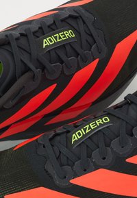 Baskets de sport noires avec des accents rouges, présentant une tige en mesh, des superpositions texturées et un logo vert sur la languette. Les lacets sont bien attachés.