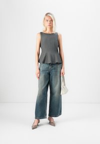Grå peplumtop med strukturerad silhuett, parat med vida blekfärgade jeans. Modellen håller en ljusgrå handväska och bär spetsiga beige skor.