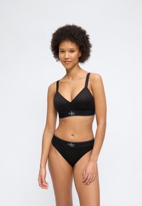 Γυναίκα με σγουρά μαλλιά, φορώντας μαύρο σουτιέν και εσώρουχα Calvin Klein, όρθια μπροστά σε απλό φωτεινό φόντο.