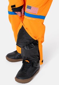 Pantaloni impermeabili di un arancione brillante con pannello inferiore nero, striscia blu e toppa con la bandiera americana, dotati di cerniere e polsini elasticizzati per una vestibilità regolabile.