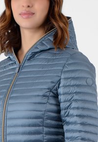 Giacca imbottita azzurro chiaro con trapuntatura orizzontale, chiusura con zip e cappuccio. Presenta un logo circolare sulla spalla sinistra. Texture liscia.