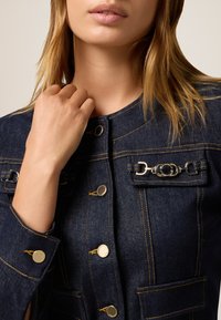 Tumman denim-takkia, jossa on hopeanväriset yksityiskohdat, koristeellinen tasku ketjuviihelyksellä, nappikiinnitys ja kontrastiväriset ompelu- ja yksityiskohtia.