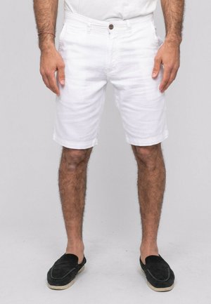 Homme portant un short blanc jusqu'aux genoux, des chaussures noires sans lacets et une chemise blanche, debout devant un fond uni.