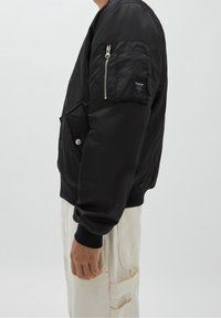 Veste bomber noire avec une poche zippée sur la manche gauche, fermeture à pression, poignets côtelés et matériau lisse. Portée avec un pantalon de couleur claire.