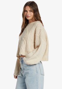 Pull en tricot crème au design court, avec des motifs en relief et des manches longues. Associé à un jean en denim bleu clair.
