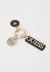 Sleutelhanger met goudkleurige hardware, een zwart leren label met een reliëf "GUESS," en een ronde bedel met een driehoekige uitsnede.