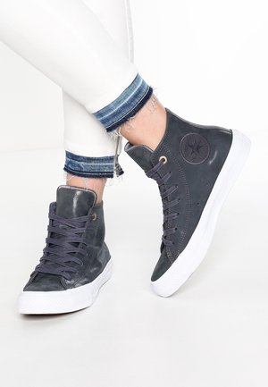 Personne portant des baskets montantes gris foncé avec des semelles blanches et des lacets foncés, associées à un pantalon blanc retroussé sur des revers en denim effiloché.
