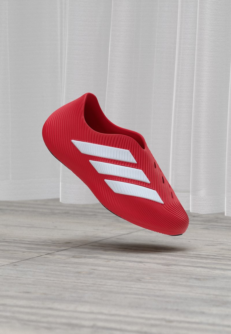 Chaussure rouge à enfiler avec un motif trois bandes blanches flottant au-dessus d’un sol en bois clair devant des rideaux blancs.