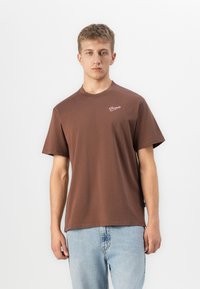 Young man wearing a brown short-sleeve T-shirt with "Pompeii" text on chest and light blue jeans, standing against a white background.