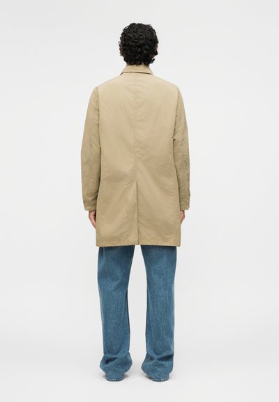 Άνδρας με σκούρα σγουρά μαλλιά, ντυμένος με μπεζ μακρύ παλτό και άνετο μπλε τζιν, στέκεται με την πλάτη προς το μέρος μας μπροστά σε ένα απλό φόντο.