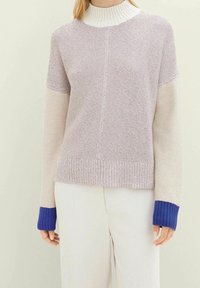Frau in einem locker gestrickten Pullover mit lila Körper, cremefarbenen Ärmeln und dunkelblauen Bündchen, kombiniert mit schneeweißen Hosen.