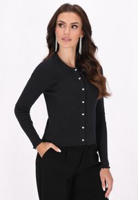Cardigan nero a costine con maniche lunghe, caratterizzato da bottoni argentati a forma di cuore e un sottile luccichio. Vestibilità slim con polsini scallop.
