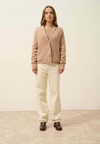 Cárdigan de punto beige con cuello en V, bolsillos y botones frontales, combinado con pantalones de pierna ancha color crema y zapatos de tacón marrón oscuro.