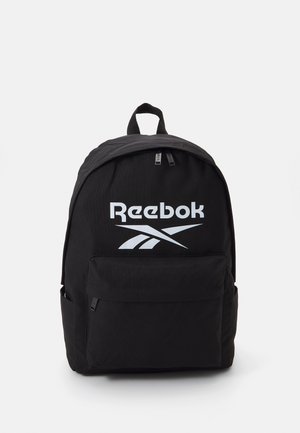 Mochila preta com uma textura suave, apresentando um grande logótipo da Reebok em branco, fecho de zíper duplo e um bolso frontal.