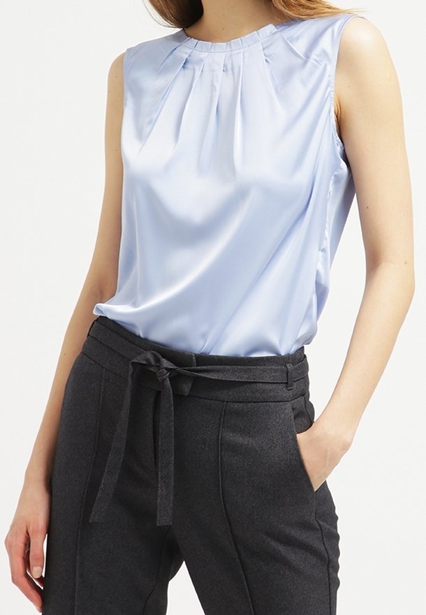 Femme portant un chemisier sans manches en satin bleu clair rentré dans un pantalon taille haute gris foncé avec une ceinture en tissu nouée à la taille.