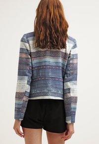 Veste tissée avec diverses nuances et motifs bleus, dotée de manches longues et d'une coupe courte. Short noir visible en bas.