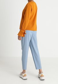 Oranssi pitkähihainen pusero, jossa on kerättyjä hihansuita, yhdistettynä vaaleansinisiin korkeasecotekoruihin ja valkoisiin lenkkikenkiin, joissa on harmaita ja keltaisia yksityiskohtia.