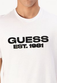 Tricou alb din bumbac, având inscripționat în negru, cu broderie îndrăzneață, textul "GUESS" și "EST. 1981". Cu guler rotund și mâneci scurte. Design simplu, fără răsfățuri.
