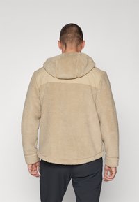 Beige Fleece-Jacke mit Kapuze, mit einem glatten, braunen Paneel über den Schultern. Weiche Textur und runder Saum.