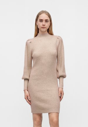 Kvinde med glat blondt hår iført en beige ribstrikket kjole med pufærmer og høj hals, stående mod en ensfarvet baggrund.