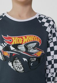 Tmavě modré tričko s dlouhým rukávem s červenou a žlutou grafikou "Hot Wheels" znázorňující závodní auto, s rukávy s kostkovaným vzorem.