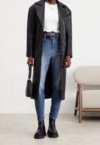 Černý kožený dlouhý kabát přes bílý krátký texturovaný top, spárovaný se světlými modrými džíny a černými robustními botami, držící malou černou kabelku.