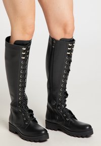 Botine negre din piele, până la genunchi, cu talpă groasă, cu șireturi în față și fermoare pe lateral, purtate de o persoană cu picioarele goale pe un fundal simplu.