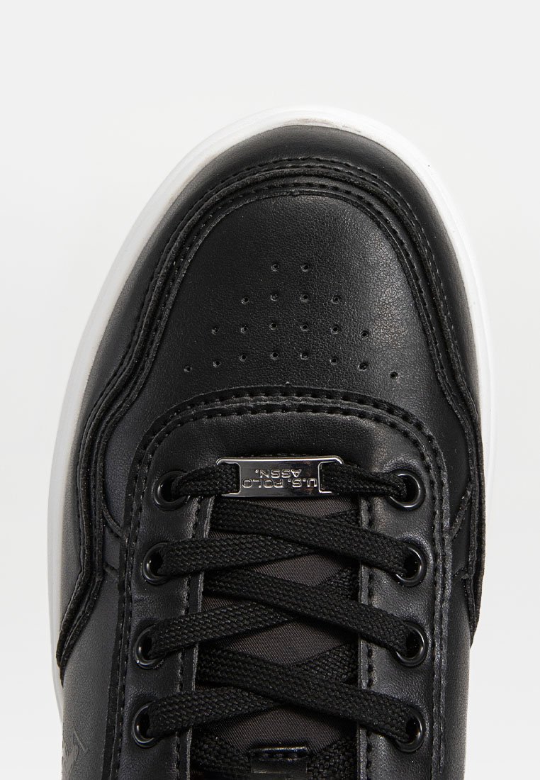 Sneaker in pelle nera con punta perforata, lacci neri, suola in gomma bianca e piastra con logo in metallo vicino ai lacci. Superfici lisce e testurizzate.