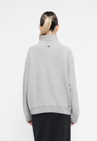 Pessoa com um rabo de cavalo loiro, vestindo um sweat de gola alta cinza com o logótipo da Adidas nas costas, combinado com uma saia preta.