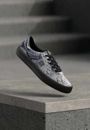 Basket avec semelle noire et lacets, motif peau de serpent bleu-gris, flottant au-dessus d'un rebord en béton avec un escalier gris flou en arrière-plan.