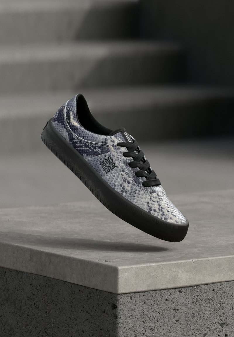 Basket avec semelle noire et lacets, motif peau de serpent bleu-gris, flottant au-dessus d'un rebord en béton avec un escalier gris flou en arrière-plan.