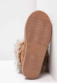 Zapatilla de ante marrón con suela de goma texturizada, con un borde superior adornado con un suave y esponjoso pelo sintético marrón claro. Talla marcada como 37.