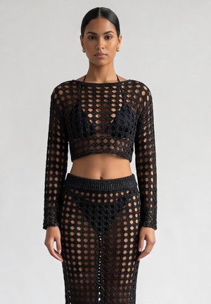 Vrouw met steil zwart haar draagt een zwarte gehaakte crop top met lange mouwen en een bijpassende rok met zwart ondergoed, staand tegen een effen achtergrond.