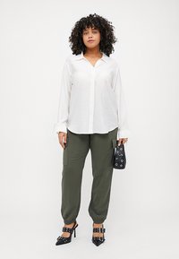 Λευκό πουκάμισο με μακριά μανίκια και κουμπιά, ελαιόχρωμο παντελόνι cargo με ελαστικές μανσέτες, μαύρες τακούνιες και μια μικρή μαύρη τσάντα με τρύπες.