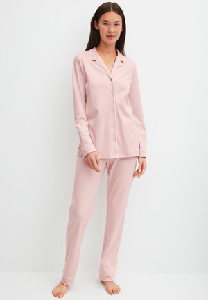 Frau steht barfuß und trägt ein hellrosa langärmeliges, geknöpftes Pyjama-Oberteil und passende Hosen mit weißer Paspel an den Bündchen und am Kragen.