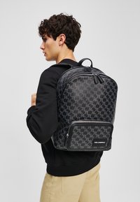 Rucsac negru din material sintetic texturat, cu un model repetitiv, fermoare duble, buzunar frontal și mâner superior.
