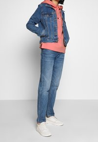 Veste en denim, sweat à capuche rose et jeans bleus à coupe décontractée. Des baskets blanches complètent la tenue. La veste dispose de poches classiques.