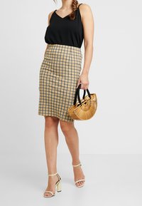 Top negro de cuello en V, falda a cuadros amarilla y azul, sandalias de tacón cuadrado beiges y un bolso circular de bambú trenzado con asas negras.