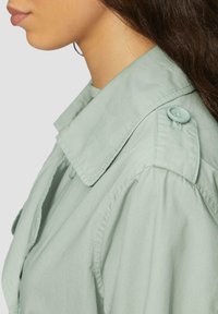 Camisa verde claro con un gran cuello puntiagudo, que presenta detalles de botones en el hombro y una textura suave y lisa.