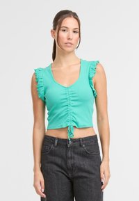 Turkusowy krótki top z falbanami na rękawach, z przodu marszczony i z wiązaniem na dole. Połączony z ciemnoszarymi dżinsami.