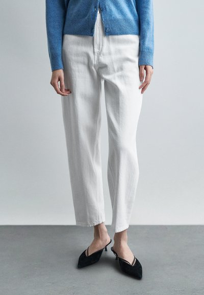 Personne portant un jean blanc taille haute, des mules à talons pointus noires et un cardigan bleu boutonné, debout sur un sol gris contre un mur gris clair.