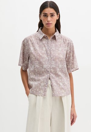 Žena oblečená v svetlofialovej kvetinovej košeli s krátkym rukávom na gombíky a krémových širokých nohaviciach, stojaca s rukami v vreckách.