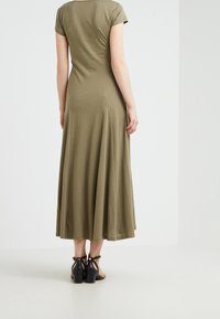 Abito verde oliva, al ginocchio, realizzato in tessuto morbido ed elastico, con maniche a cappuccio e gonna svasata a forma di A. Indossato con sandali neri con tacco.