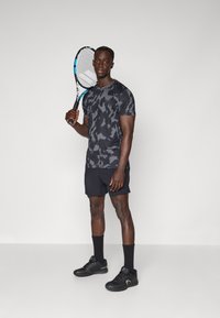 T-shirt de sport à motif camouflage noir et gris avec des manches courtes, associé à un short noir. Tenant une raquette de tennis, portant des chaussures de sport noires.