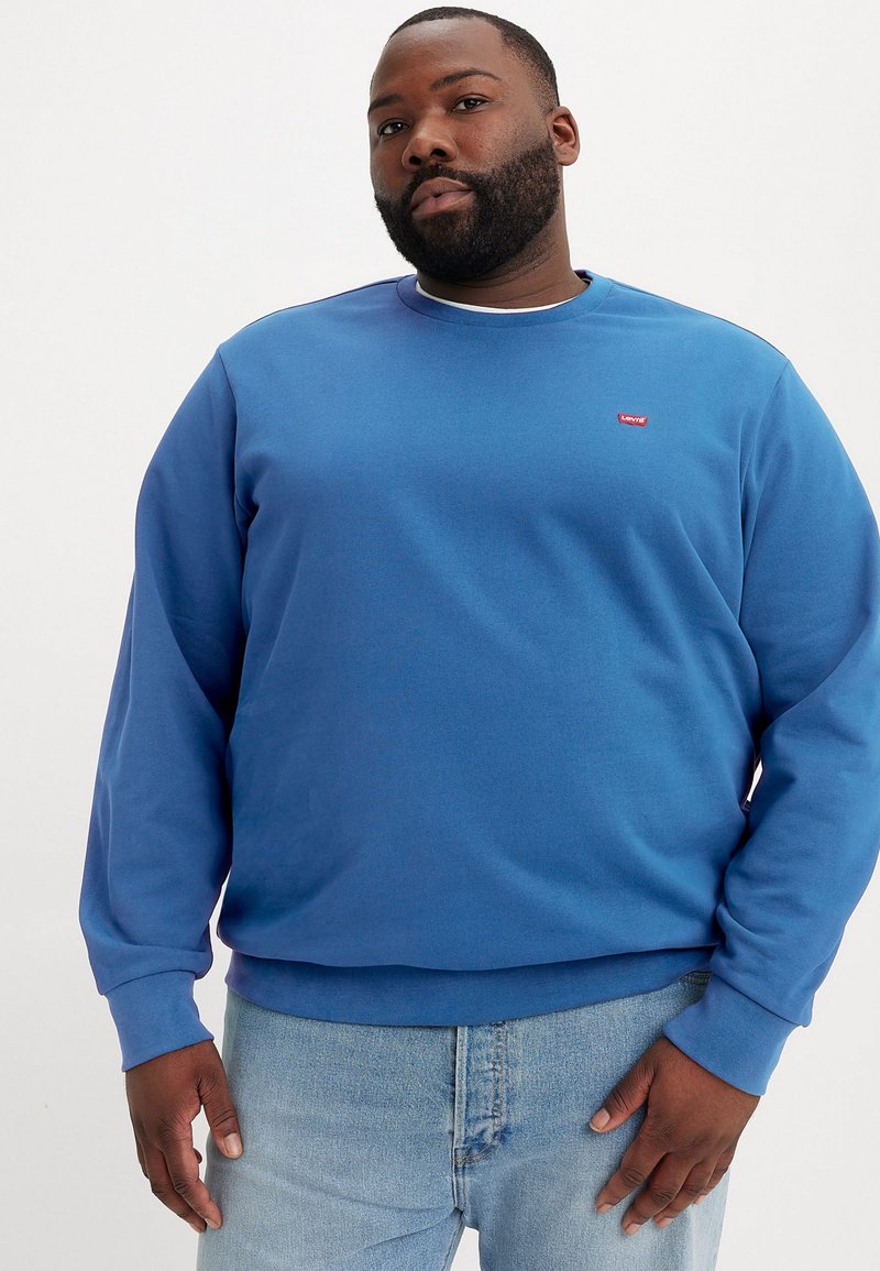Blue sweatshirt with a ribbed crew neckline and long sleeves. Features a small red logo on the chest. Paired with light-wash denim jeans.